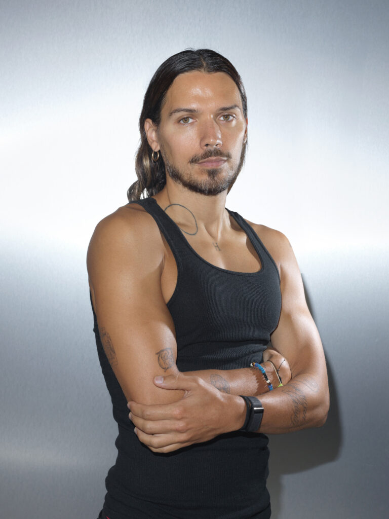 A person with tattoos wearing a black tank top poses with arms crossed against a plain background.